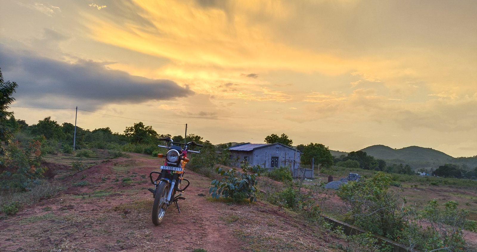 A motorcycle standing alone on a red earth track along a ridge, a grey plastered house with a metal roof behind it, hills and a yellow evening sky