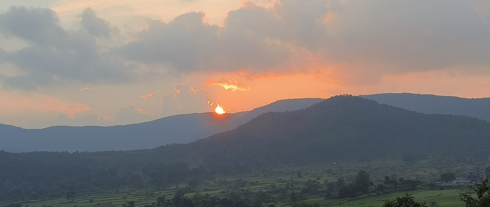 The sun setting behind a forested ridge, orange cloud above it, terraced fields and scattered trees on the slope below