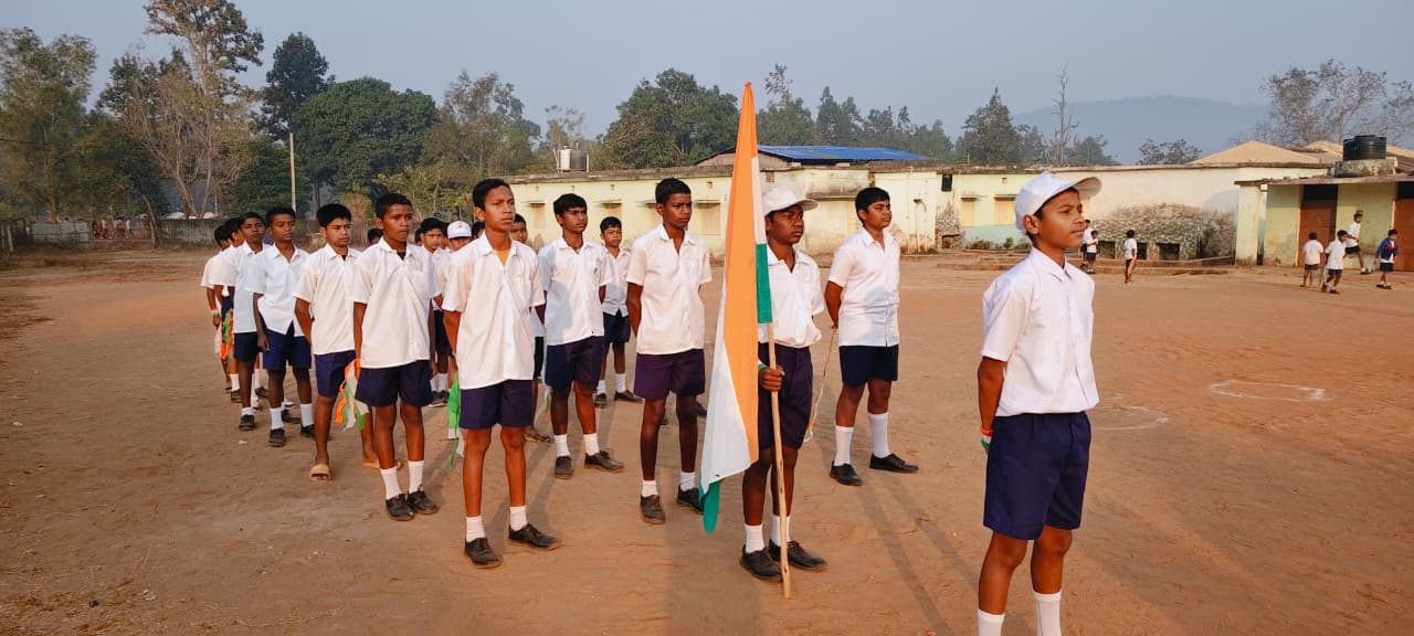 Rows of boys in white shirts and navy shorts standing in formation on a red earth ground, one holding the national flag