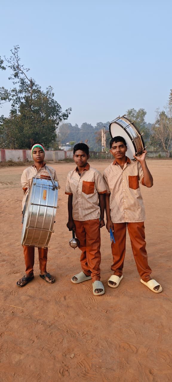 Three older students on red earth holding a dhol and a snare drum, one wearing a tricolour headband