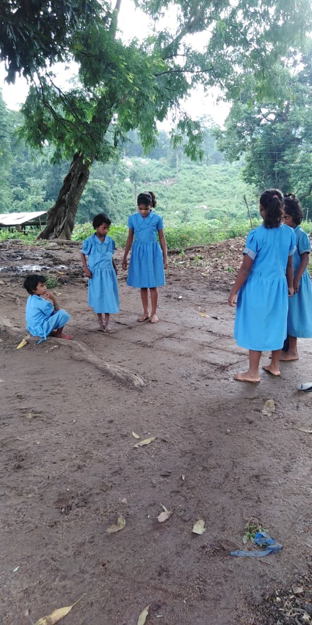 Six girls in blue school uniforms standing barefoot on wet earth under a large tree