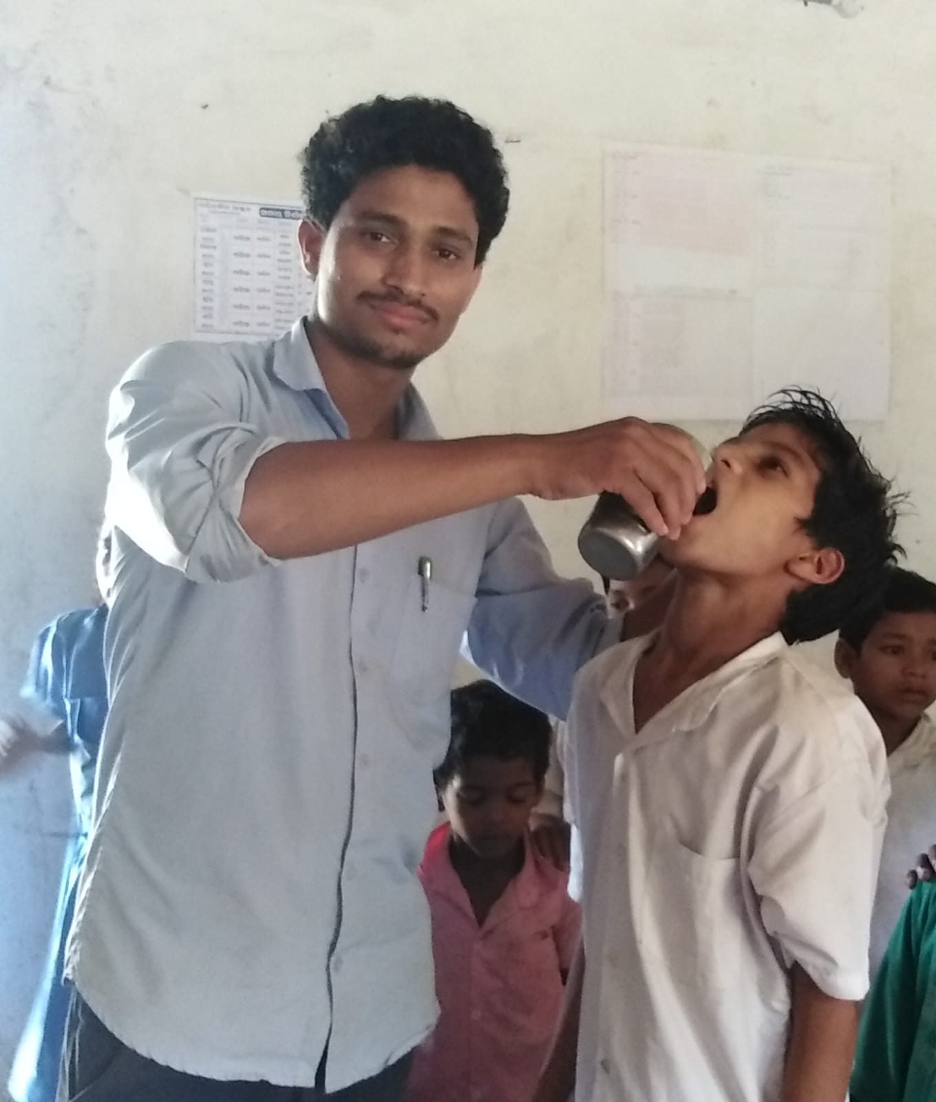 A man pouring from a steel tumbler into a boy's mouth in a classroom, other children waiting