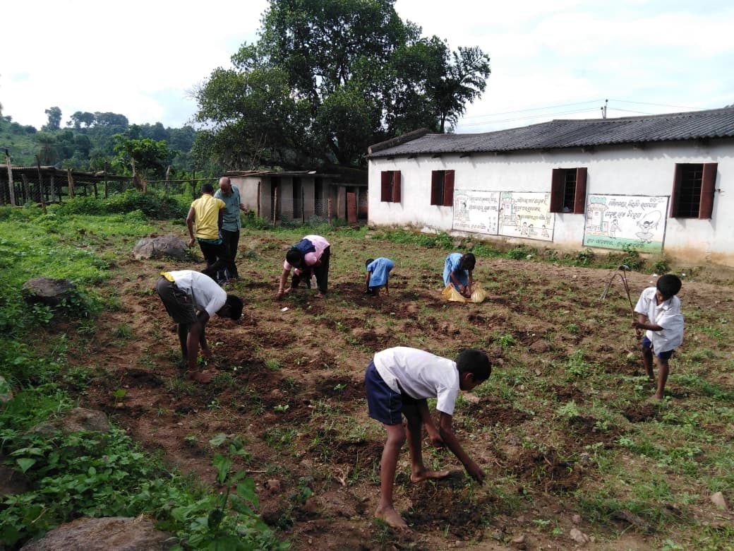 Children and two adults bent over a tilled plot beside a school building painted with Odia murals