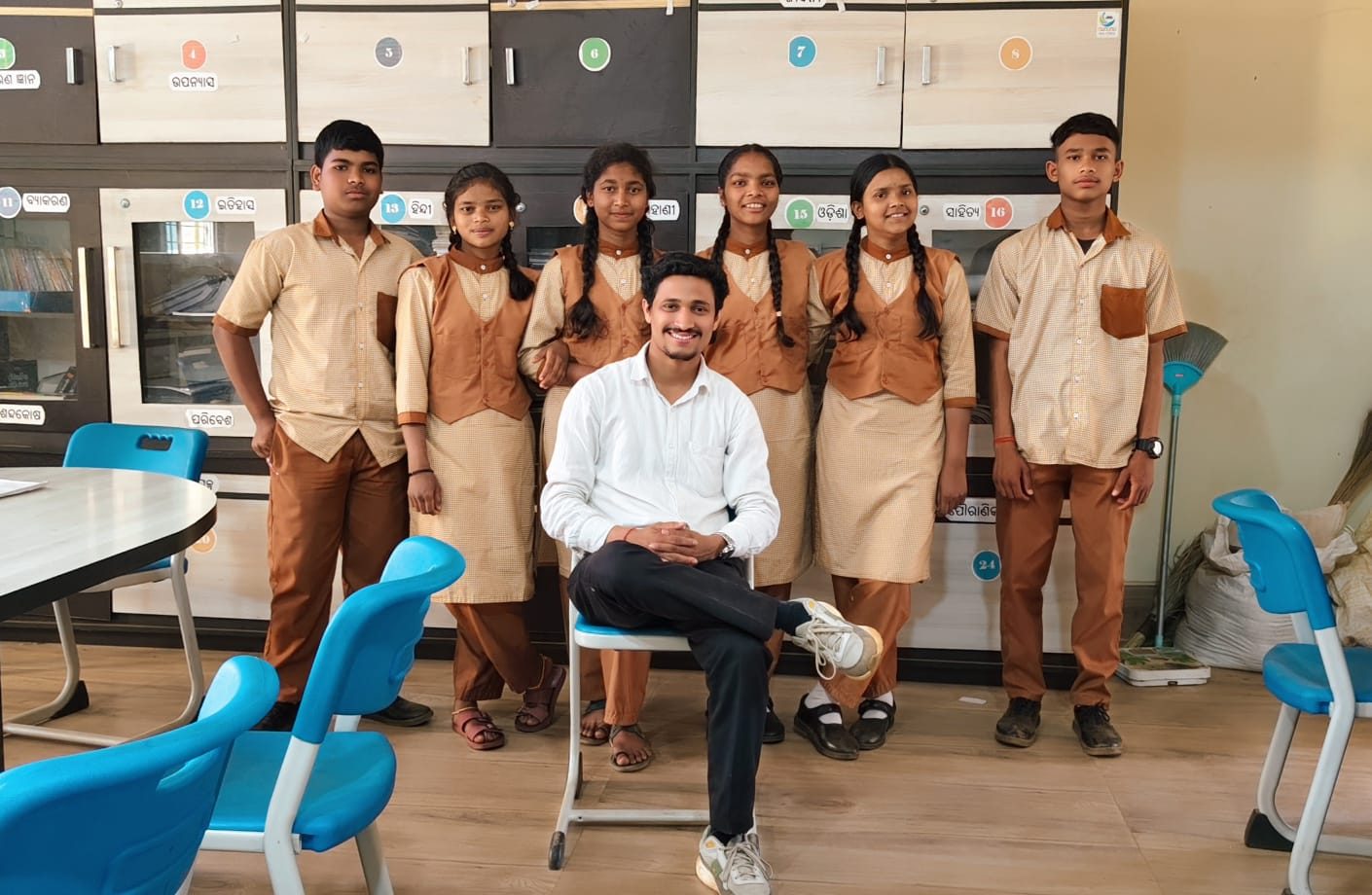 Six older students standing behind a seated young man in a school library lined with numbered subject cupboards