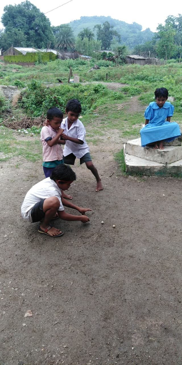 Three boys crouched over marbles on packed earth, a girl in a blue uniform watching from concrete steps
