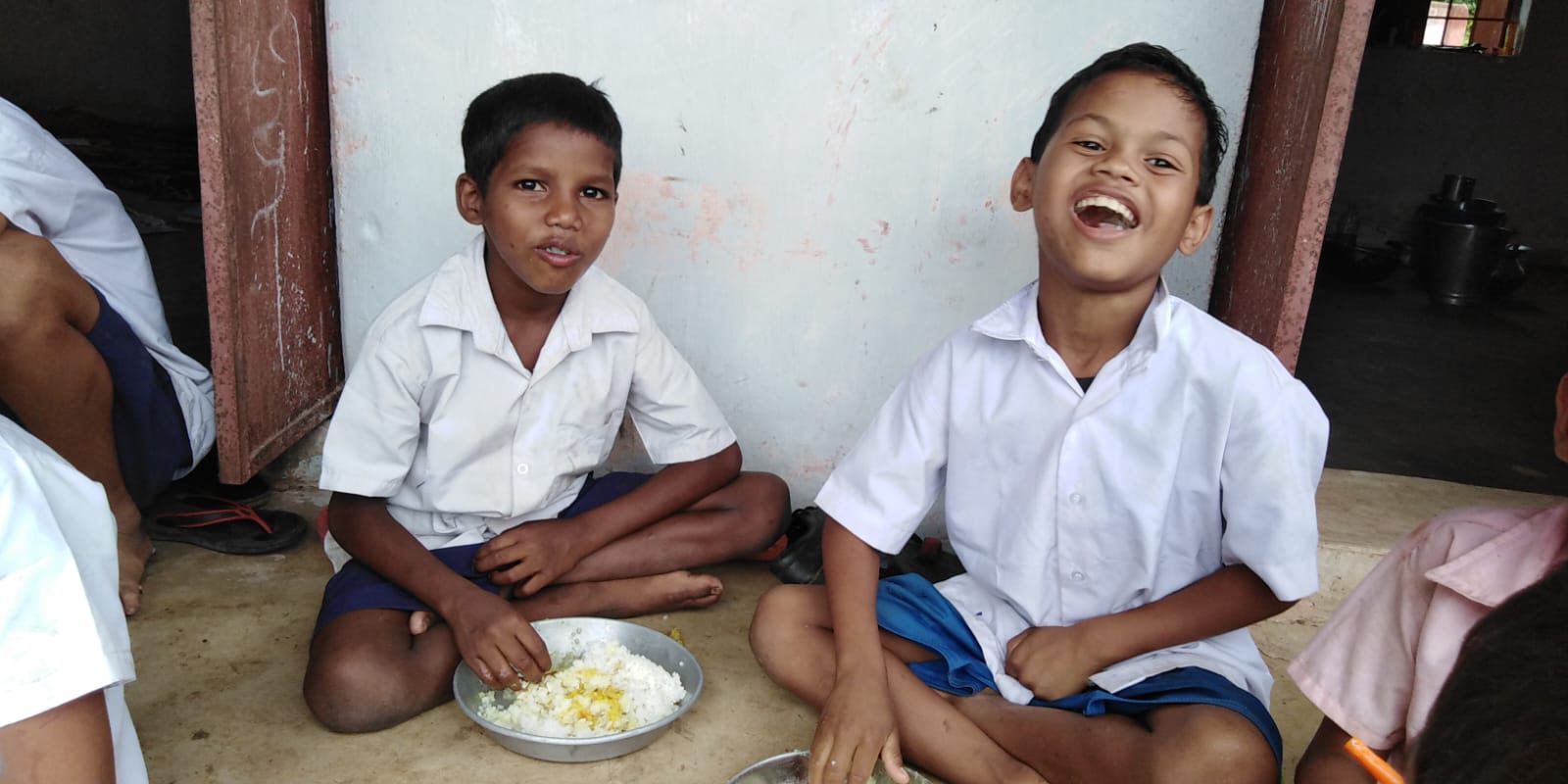 Two boys in school uniform sitting on the floor eating rice from steel plates, one laughing