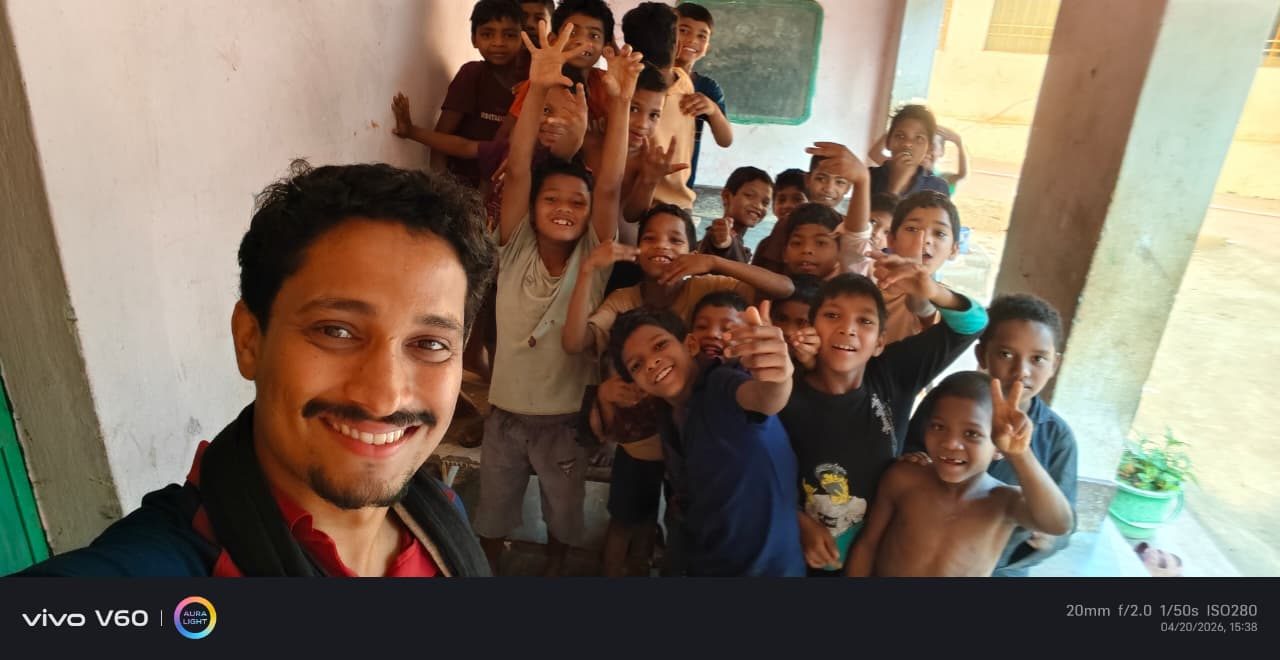 A man taking a selfie with about fifteen children crowding and waving behind him in a school veranda