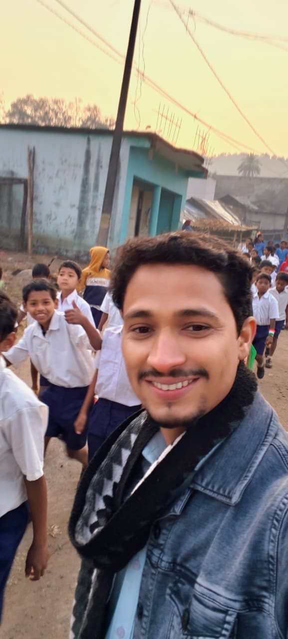 A man taking a selfie on a village road at dusk with schoolchildren in uniform walking behind him