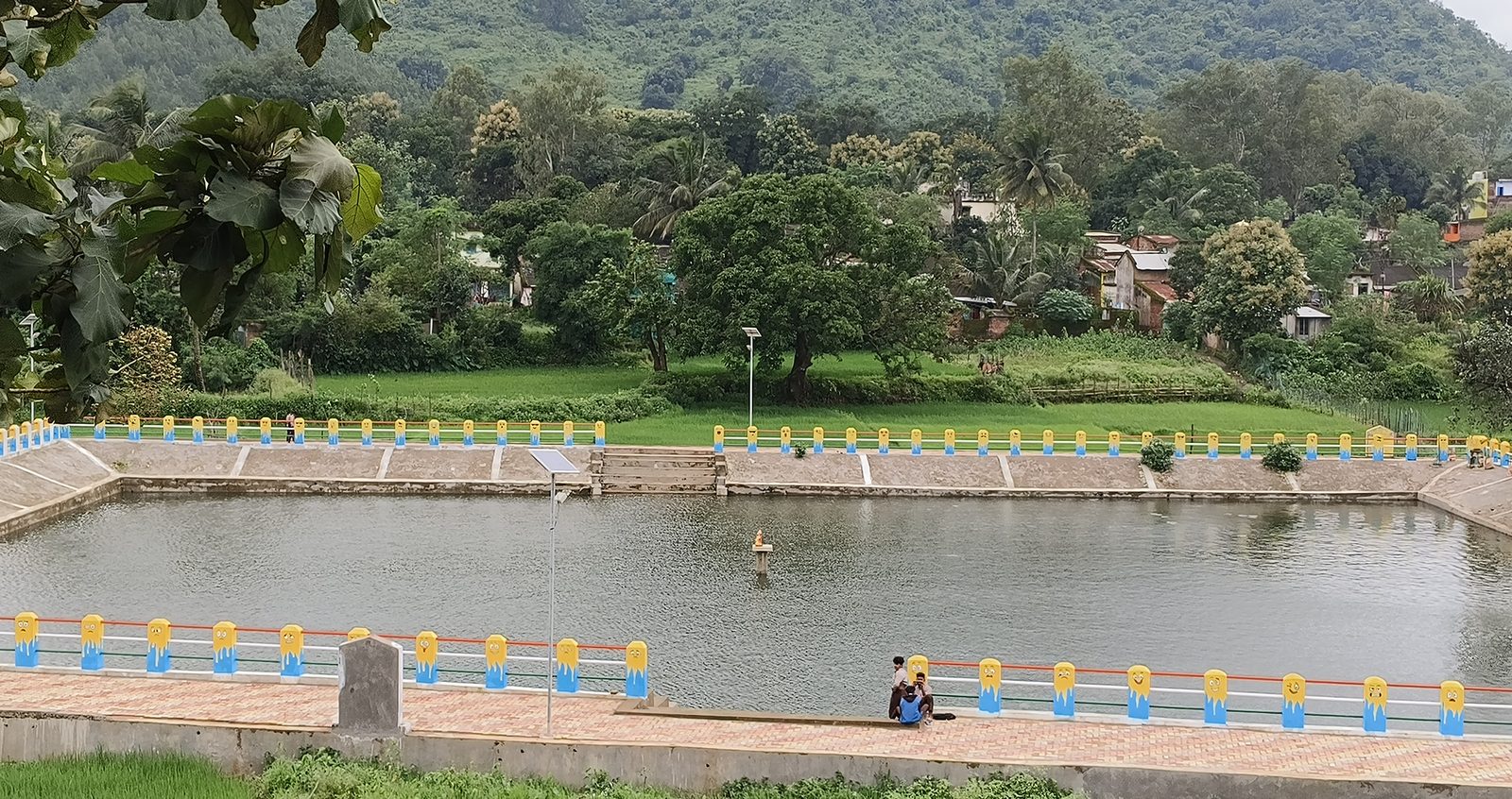 A rectangular village tank with stepped concrete sides, the railings around it painted yellow and blue, green paddy and houses on the far bank with a hill behind